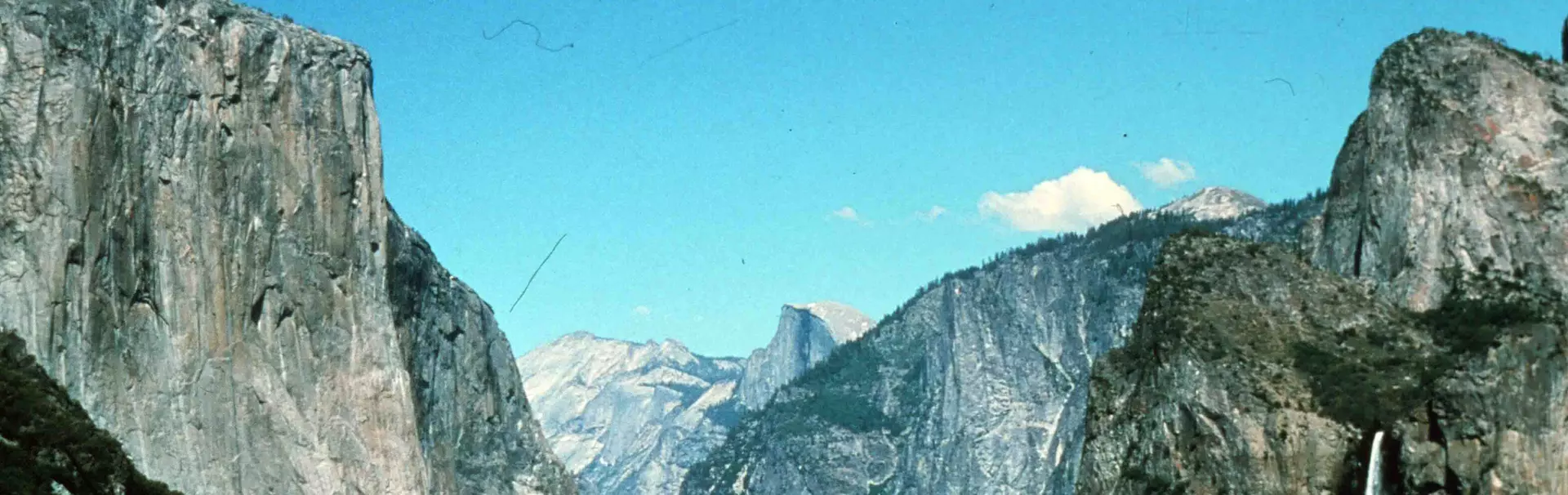 Een schilderachtig uitzicht op de Yosemite-vallei met hoge granieten rotswanden, een beboste vallei en bergen in de verte onder een helderblauwe hemel. Aan de rechterkant van de foto is een waterval t